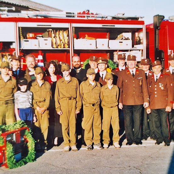Eine Gruppe von Personen in Militäruniformen steht vor einem Feuerwehrwagen. Einige tragen Mützen, und auf den Uniformen sind Medaillen sichtbar.