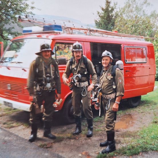Drei Feuerwehrleute in Uniform stehen vor einem roten Feuerwehrwagen mit einer Leiter oben drauf. Sie lächeln und sehen bereit für den Einsatz aus.