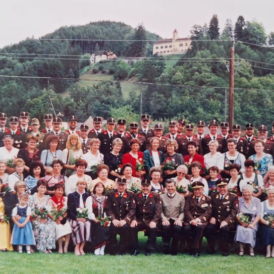 Eine Gruppenaufnahme von uniformierten Offizieren und Zivilisten, einschließlich Kindern, die auf einem Rasenfeld stehen und sitzen, mit einem Hintergrund aus Bäumen und einem Gebäude.