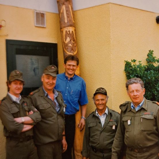 Fünf Männer in Militäruniformen und ein Mann in einem blauen Hemd posieren für ein Foto vor einem Gebäude.