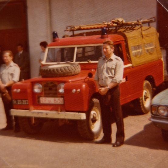 Ein Vintage-Foto eines Mannes, der vor einem roten Feuerwehrauto mit einem Ersatzreifen auf dem Dach steht.