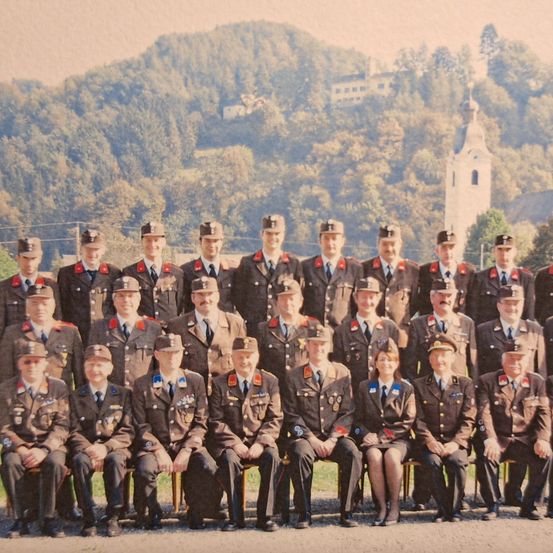 Eine Gruppe von Militärangehörigen in Uniformen posiert für ein Foto vor einem Hügel mit Bäumen und einem Turm im Hintergrund.