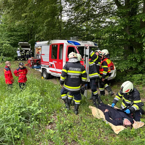 Feuerwehrleute in voller Montur kümmern sich um eine verletzte Person auf einer Trage in einem bewaldeten Gebiet. Die Szene befindet sich in der Nähe eines Feuerwehrwagens mit offener Tür, mit weiterem Personal in der Nähe.