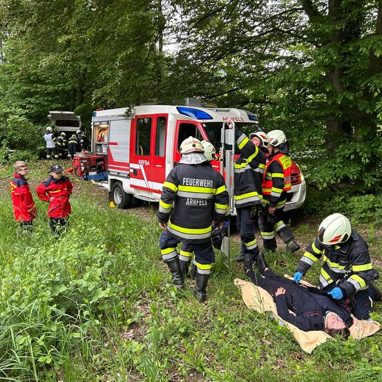Feuerwehrleute in voller Montur kümmern sich um eine verletzte Person auf einer Trage in einem bewaldeten Gebiet. Die Szene befindet sich in der Nähe eines Feuerwehrwagens mit offener Tür, mit weiterem Personal in der Nähe.