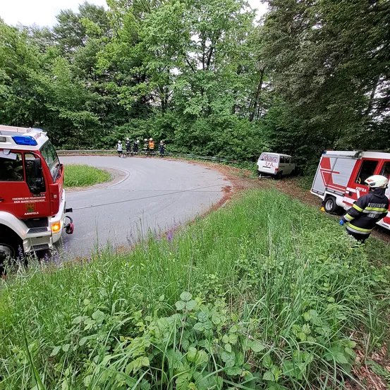 Feuerwehrleute in Schutzausrüstung stehen am Straßenrand. Zwei Feuerwehrwagen sind geparkt. Ein weißer Kleintransporter steht am Grasstreifen. Menschen in Warnwesten sind in der Nähe des Kleintransporters.