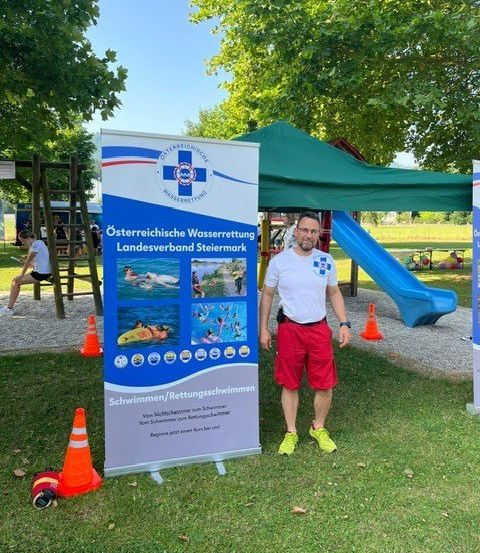 Ein Mann steht neben einem Banner zur Wassersicherheit in einem Park, mit einer blauen Rutsche und einem Zelt im Hintergrund.