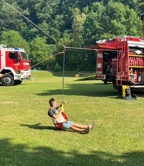 Ein junger Junge genießt eine Fahrt auf einer Schaukel, die an einem Feuerwehrauto befestigt ist, in einem Park. In der Nähe ist ein weiteres Feuerwehrauto geparkt.