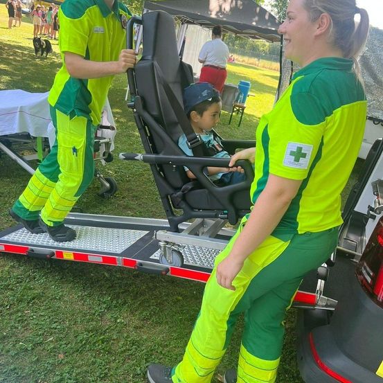 Zwei Personen in grünen Uniformen entladen an einem sonnigen Tag ein Kind von einer Krankenwagen-Trage.