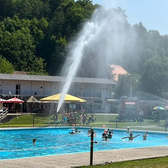 Ein Wasserbrunnen spritzt Wasser in die Luft an einem Freibad. Menschen schwimmen und einige stehen am Rand des Beckens. Im Hintergrund sind Schirme aufgestellt.
