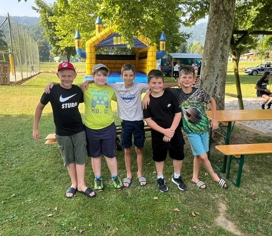 Fünf Jungen stehen auf einer Wiese in einem Park. Ein Junge trägt einen Hut. Hinter ihnen ist eine Bank und eine Hüpfburg. In der Ferne ist ein Polizeiauto geparkt.