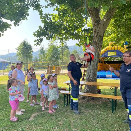 Eine Gruppe von Kindern und Erwachsenen versammelt sich in einem Park. Ein junges Mädchen schwingt sich an einem Baum hoch. Zwei Männer halten Seile für sie. Sie stehen vor einem Picknicktisch und einem Hüpfburg. Im Hintergrund befinden sich ein Zaun, Bäume und Berge.