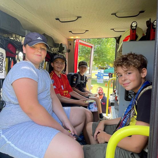 Vier Kinder sitzen lächelnd in einem Feuerwehrauto. Eines trägt eine graue Adidas-Kappe. Die anderen tragen rote, schwarze und gelbe T-Shirts. Ein erwachsener Mann ist im Hintergrund.