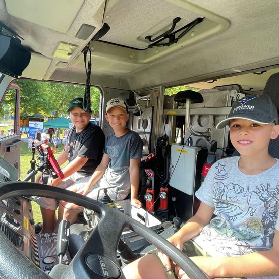 Drei Jungen sitzen lächelnd in einem Feuerwehrauto. Der vordere trägt ein graues T-Shirt und eine blaue Baseballkappe. Hinter ihm tragen zwei andere Jungen grüne Mützen. Das Innere des Fahrzeugs ist mit Ausrüstung ausgestattet, und draußen ist ein sonniger Tag mit Menschen zu sehen.