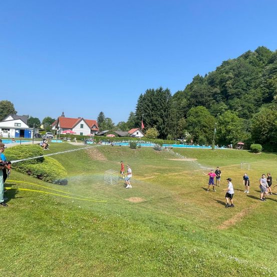 Eine Gruppe von Menschen spielt ein Wasserspiel auf einem Rasenfeld, mit einem Schwimmbad und Häusern im Hintergrund. Einige tragen Hüte und Turnschuhe.