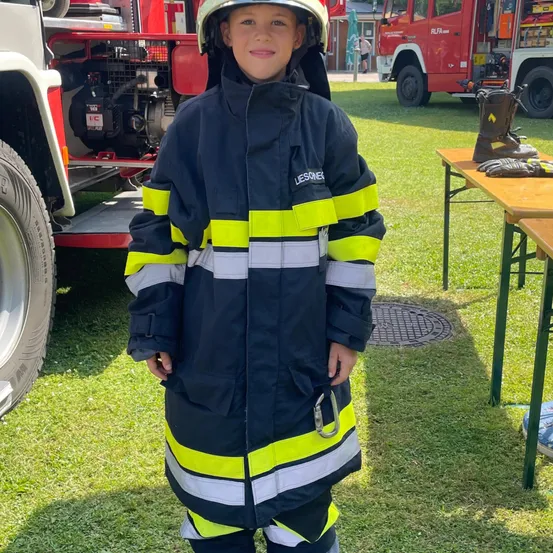 Ein junger Junge steht draußen und trägt eine Feuerwehruniform, mit einem Feuerwehrwagen im Hintergrund.