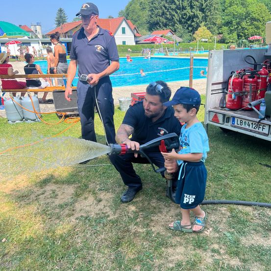 Ein junger Junge spielt mit einem Feuerlöscher, während zwei Feuerwehrleute neben ihm in der Nähe eines Schwimmbeckens stehen.
