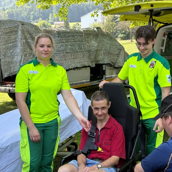 Zwei medizinische Helfer unterstützen einen Mann im Rollstuhl, der Sicherheitsgurte trägt. Sie stehen neben einem Krankenwagen, mit einem abgedeckten Fahrzeug und einem weißen Gebäude im Hintergrund.