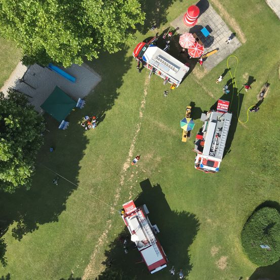 Eine Draufsicht auf einen Park mit einem Feuerwehrauto, einem Eiswagen und einigen Personen. Es gibt auch Bäume, ein Zelt und eine Rutsche.