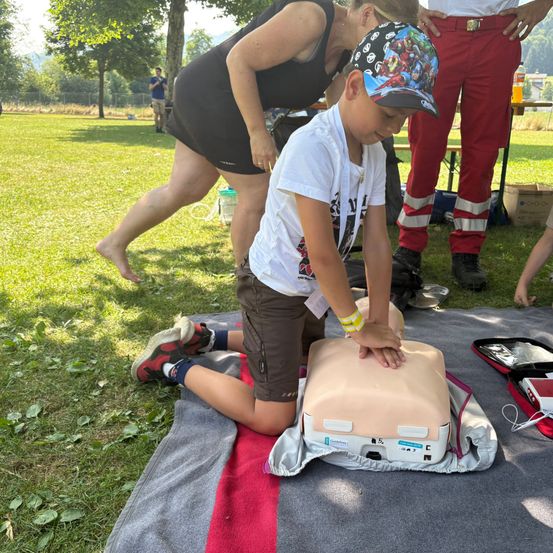 Ein junger Junge übt CPR an einem Dummies auf einer Decke in einem Park. Eine Frau und ein Mann in roten Hosen beobachten.