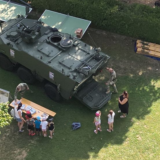Eine Luftaufnahme eines Militärfahrzeugs mit Soldaten und Kindern. Eine Frau und Kinder stehen neben einem Tisch mit Gegenständen darauf.