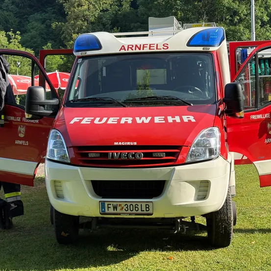 Ein roter und weißer Feuerwehrwagen aus Arnfels steht mit offenen Türen auf dem Rasen. Das Wort 'Feuerwehr' ist auf der Front und 'Magirus' auf dem Stoßfänger zu sehen.