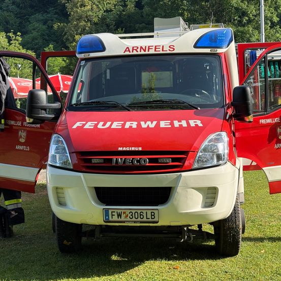 Ein roter und weißer Feuerwehrwagen aus Arnfels steht mit offenen Türen auf dem Rasen. Das Wort 'Feuerwehr' ist auf der Front und 'Magirus' auf dem Stoßfänger zu sehen.
