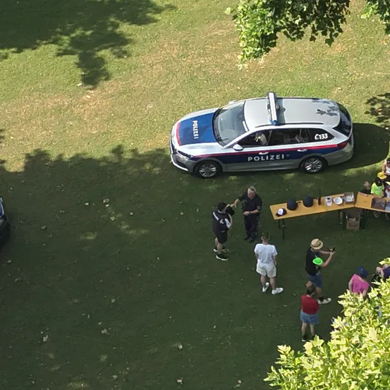 Eine Vogelperspektive zeigt einen Polizeiwagen, der auf einem Rasen geparkt ist. In der Nähe versammeln sich mehrere Personen, eine hält eine Kamera. Ein Tisch mit Hüten und Gegenständen ist aufgebaut.