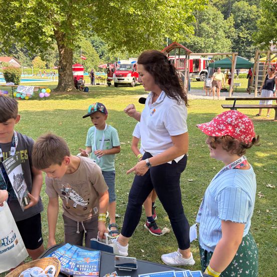 Eine Frau steht mit mehreren Kindern bei einer Outdoor-Veranstaltung, in der Nähe eines Tisches mit Broschüren, während ein Feuerwehrauto im Hintergrund geparkt ist.