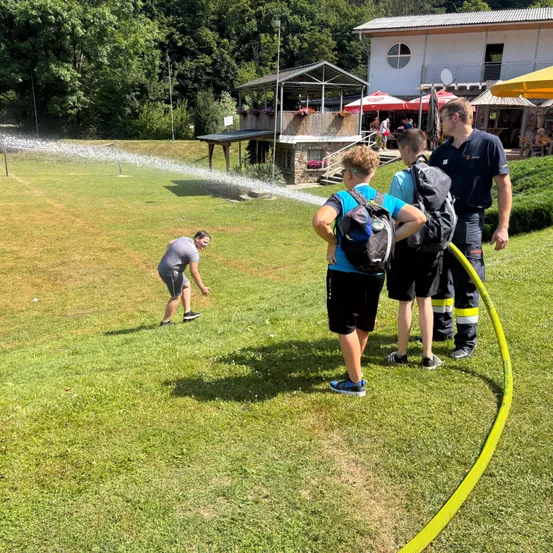 Ein Wasserschlauch sprüht Wasser auf eine Rasenfläche. In der Nähe sind vier Personen, darunter ein Feuerwehrmann und ein Junge mit Rucksack.