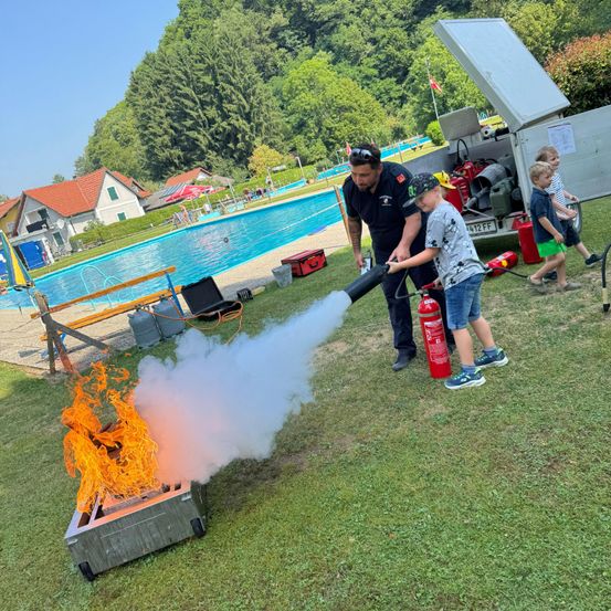 Ein Feuerwehrmann hilft einem Kind, ein kleines Feuer im Freien mit einem Feuerlöscher zu löschen. Dahinter sind ein Pool und Häuser zu sehen.