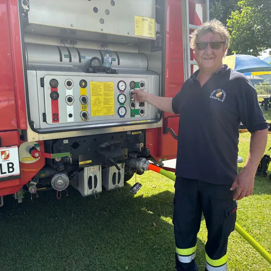 Ein Feuerwehrmann zeigt auf das Bedienfeld eines Feuerwehrwagens. Der Wagen hat einen roten Körper mit einem Nummernschild, das LB liest. Hinter ihm ist ein Grasbereich mit Zelten.