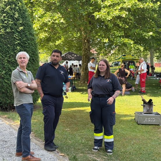 Drei Personen in Uniform stehen gemeinsam in einem Park. Dahinter sind mehrere Menschen bei Aktivitäten, einige in roten Uniformen. In der Nähe befindet sich ein Krankenwagen und ein Zelt.