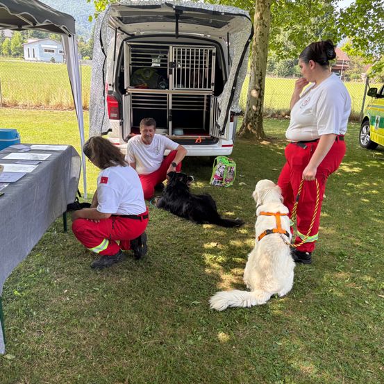 Eine Gruppe von Menschen und Hunden befindet sich auf einer Rasenfläche, zwei Hunde liegen auf dem Boden, während eine Frau eine Leine hält. Ein Mann sitzt neben ihnen auf dem Boden, mit einem Hund vor ihm. Ein Van ist hinter ihnen geparkt.