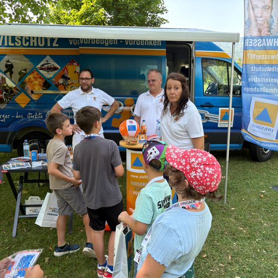 Zwei Erwachsene und vier Kinder versammeln sich um einen Tisch mit einem Tischzelt in einem Park. Ein offenes Fahrzeug zeigt Bilder von Feuer, Rettung und Wasser. Ein Schild steht für Zivilschutz vorbeugen vordenken.