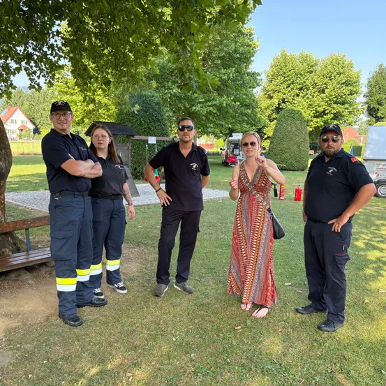Fünf Menschen stehen in einem Park und lächeln für ein Foto. Zwei Frauen und drei Männer, alle in schwarzen Uniformen, posieren vor einem Baum. Die Frau in der Mitte trägt ein Kleid und hält eine Handtasche. Hinter ihnen steht eine Bank, ein Feuerlöscher und ein Haus.