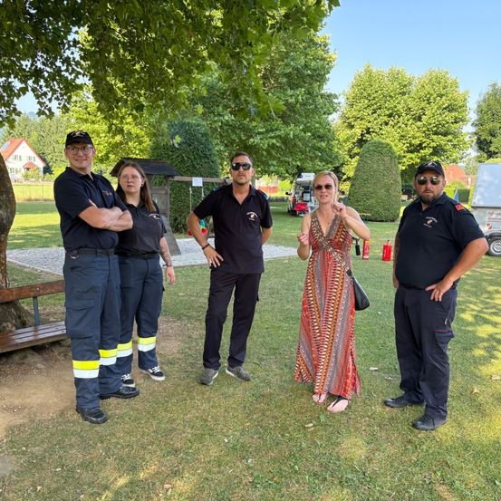 Fünf Menschen stehen in einem Park und lächeln für ein Foto. Zwei Frauen und drei Männer, alle in schwarzen Uniformen, posieren vor einem Baum. Die Frau in der Mitte trägt ein Kleid und hält eine Handtasche. Hinter ihnen steht eine Bank, ein Feuerlöscher und ein Haus.