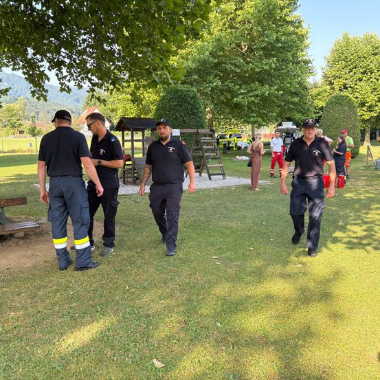 Eine Gruppe von Feuerwehrleuten in Uniform geht auf einem Rasenfeld. In der Nähe sind zwei Rettungsfahrzeuge geparkt und einige Leute stehen herum.