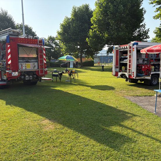Zwei Feuerwehrfahrzeuge sind auf einem Rasenfeld geparkt. Eines hat die hintere Tür offen und Werkzeuge sind im Inneren sichtbar. Ein Regenschirm steht in der Nähe. Bäume und ein Gebäude sind im Hintergrund.
