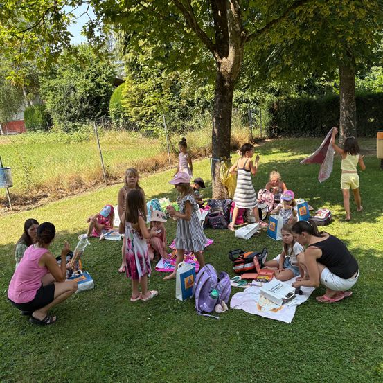 Eine Gruppe von Kindern versammelt sich auf einem Rasenfeld, möglicherweise in einem Sommercamp. Sie sind von verschiedenen Gegenständen umgeben, darunter Rucksäcke, Spielzeug und eine Picknickdecke. Einige stehen, während andere auf dem Boden sitzen. Bäume und Büsche sind im Hintergrund.