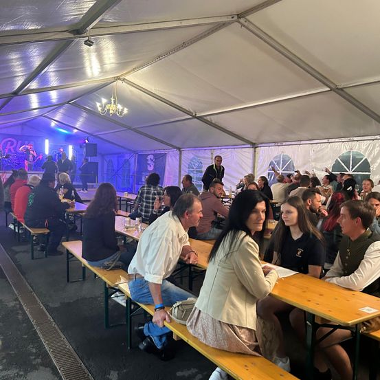 Eine Menschenmenge sitzt auf Bänken in einem Zelt. Sie essen gemeinsam. Auf der Bühne im Hintergrund spielt eine Band.