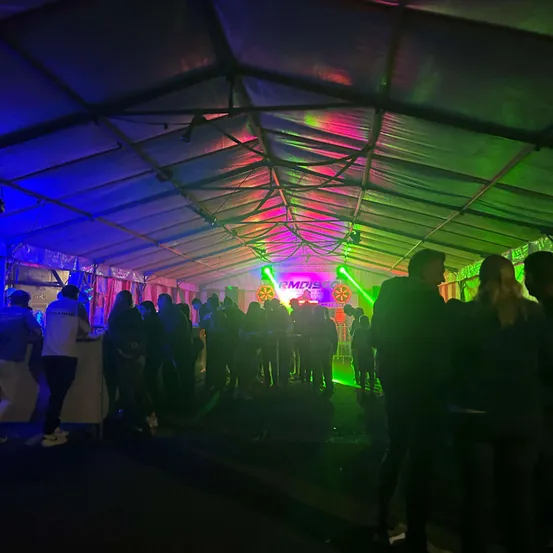 Unter einem großen Zelt versammeln sich Menschen bei Nacht mit leuchtenden, farbigen Lichtern, darunter Grün und Lila, die die Szene beleuchten. Einige Leute stehen in der Mitte, während andere an den Seiten positioniert sind.
