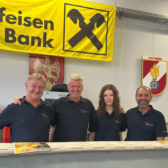 Vier Menschen stehen lächelnd für ein Foto vor einem Tresen, mit einem gelben Banner und Logos im Hintergrund.
