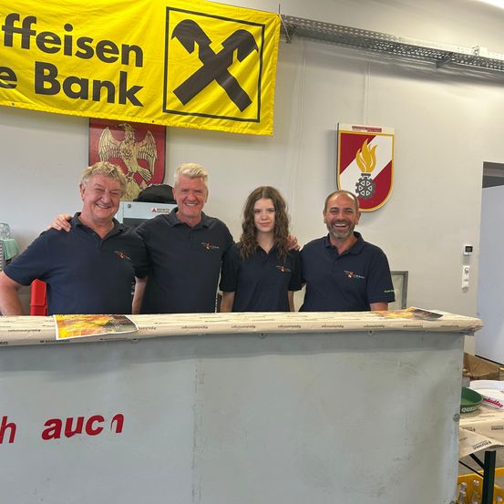 Vier Personen stehen lächelnd für ein Foto hinter einem Tresen in einem Raum. Ein gelber Banner und eine Flagge hängen an der Wand hinter ihnen.