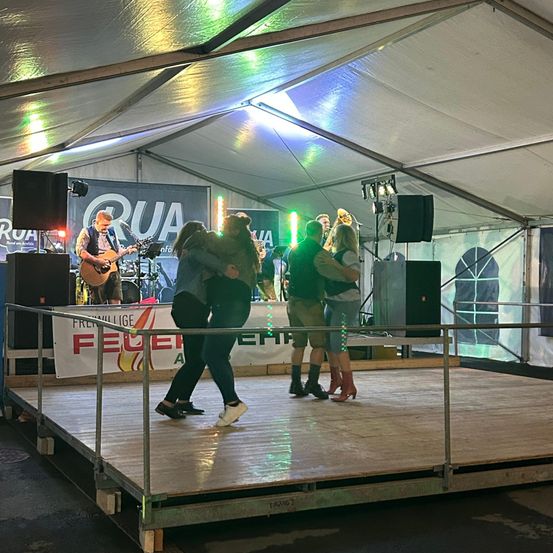 Eine Gruppe von Menschen tanzt auf einer Bühne in einem Zelt, während ein Gitarrist im Hintergrund spielt. Die Bühne ist mit Lautsprechern ausgestattet, und es gibt Banner mit Text und Logos an den Seiten.