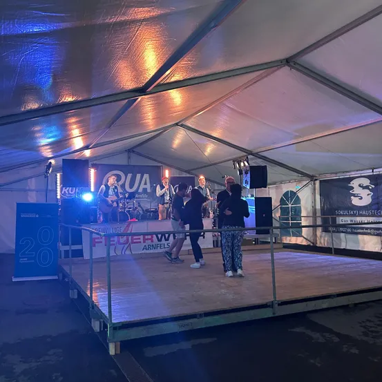 Eine Band tritt auf einer Bühne unter einem Zelt auf. Zwei Personen stehen auf der Bühne, während andere gehen. Ein Banner mit dem Wort 'Feuerwehr' befindet sich an der Seite.