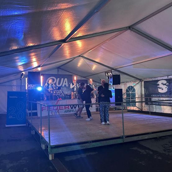 Eine Band tritt auf einer Bühne unter einem Zelt auf. Zwei Personen stehen auf der Bühne, während andere gehen. Ein Banner mit dem Wort 'Feuerwehr' befindet sich an der Seite.
