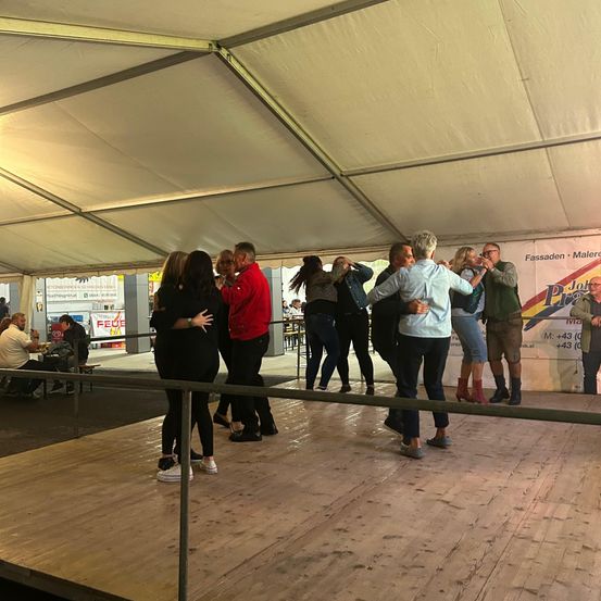 Eine Gruppe von Menschen tanzt auf einer Bühne in einem Zelt. Einige tragen Freizeitkleidung. Im Hintergrund befindet sich ein weißes Banner und Schilder.