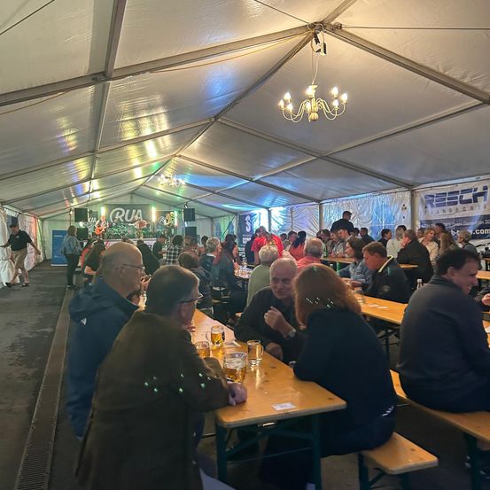 Ein Zelt mit Bühne beherbergt eine Versammlung von Menschen, die an Tischen sitzen. Getränke und Speisen werden auf den Tischen serviert. An der Decke hängt eine Kronleuchter.