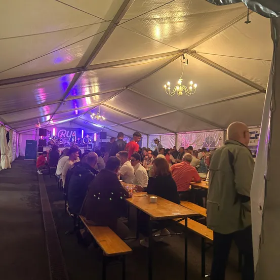 Eine große Gruppe von Menschen diniert in einem Zelt mit einer Bühne im Hintergrund. Es gibt eine Kristallleuchte und violette Beleuchtung.