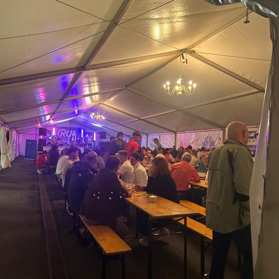 Eine große Gruppe von Menschen diniert in einem Zelt mit einer Bühne im Hintergrund. Es gibt eine Kristallleuchte und violette Beleuchtung.